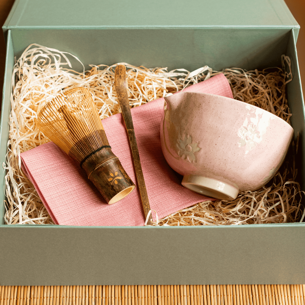Tea set with pink ceramic cup, bamboo whisk, and cloth in a green box on a wooden surface.
