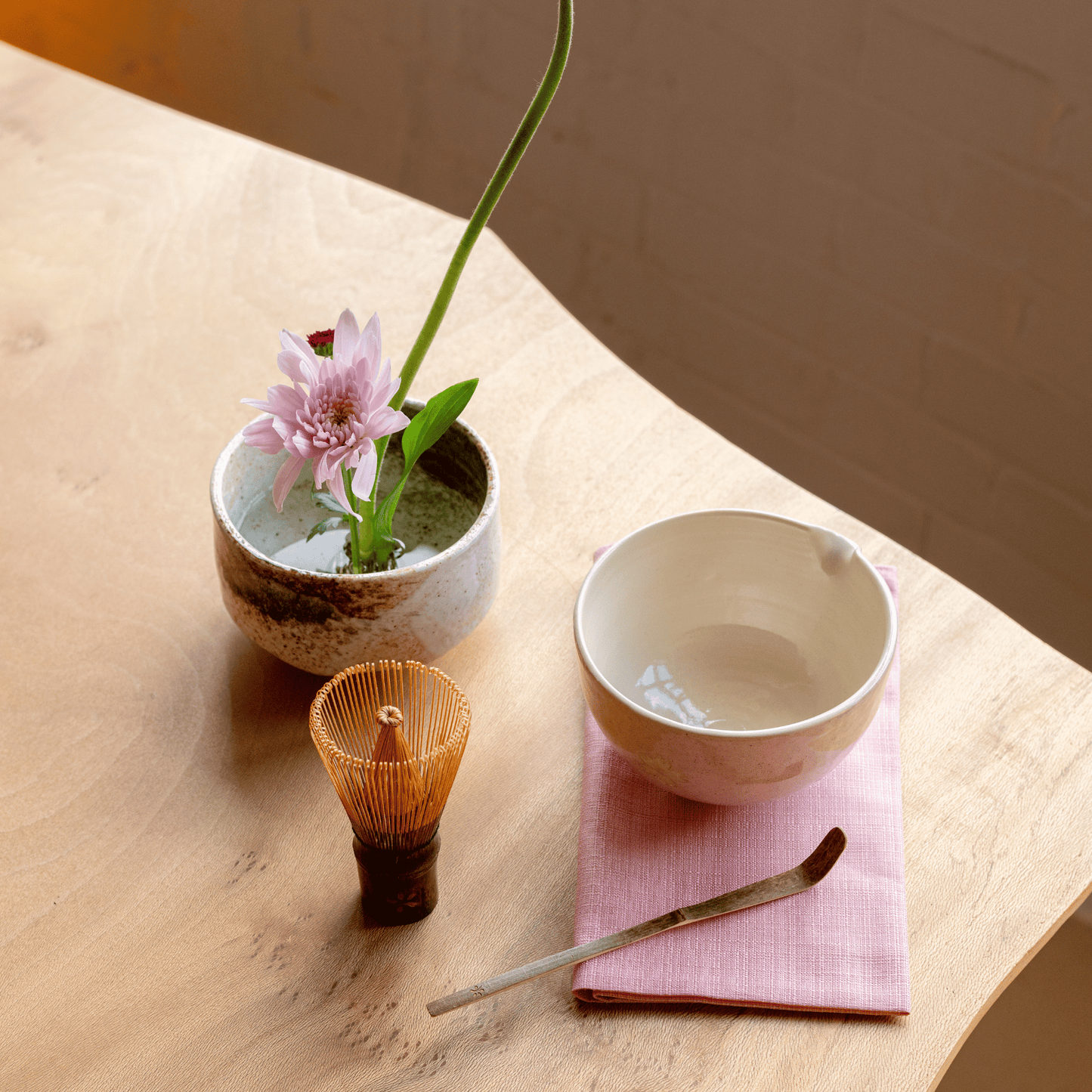 Tea-making set with a bowl, whisk, and flower on a wooden surface.
