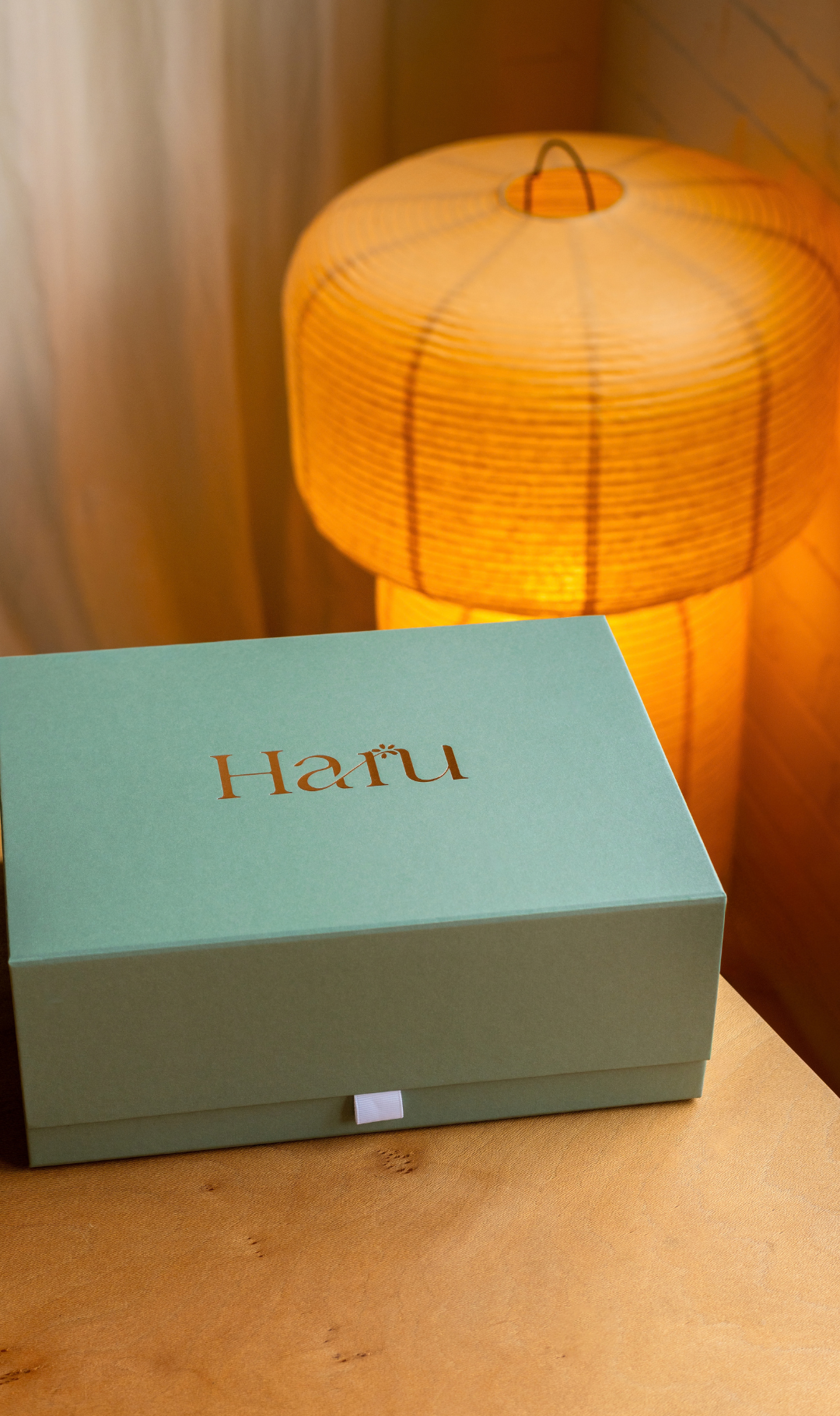 A mint green rectangular box with the brand name 'Haru' printed in gold on the lid, placed on a wooden surface in front of a glowing warm-toned paper lantern lamp.