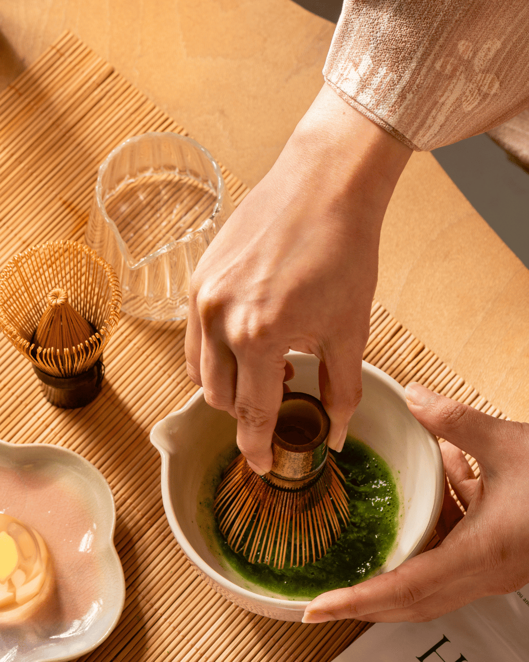 A close-up of hands whisking green matcha tea in a small white bowl using a bamboo whisk, with additional bamboo tools and a glass on a woven mat background.