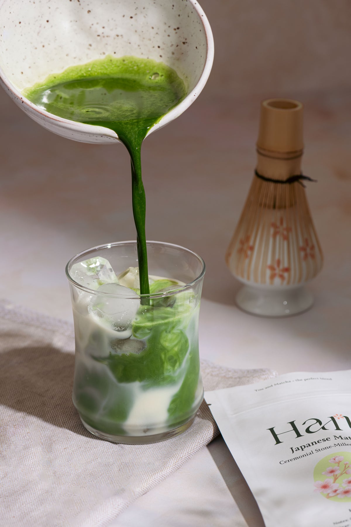 Hana Matcha being poured from a ceramic pitcher into a glass with ice cubes on a textured surface.