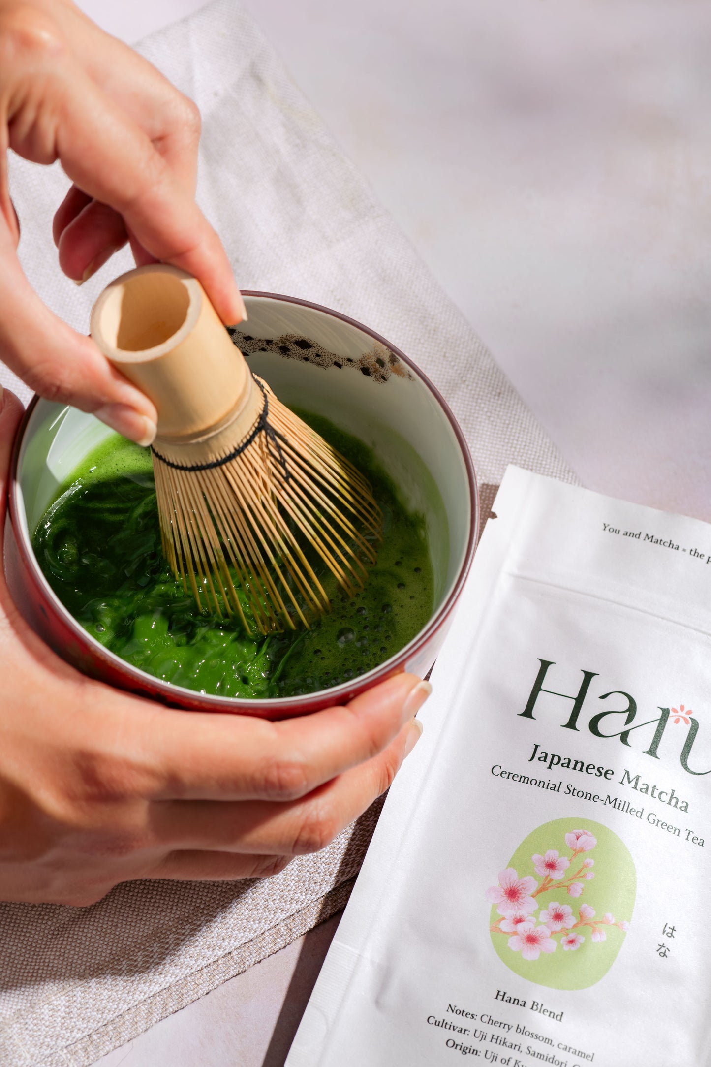 Person whisking Hana matcha green tea in a bowl with a packet of Hana Japanese Matcha next to it.