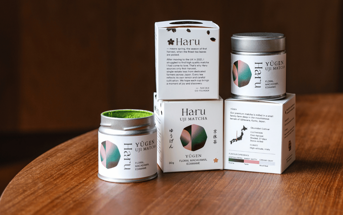 Four matcha tins from Haru including two white boxes and two silver jars, arranged on a wooden table with a blurred dark wood background.