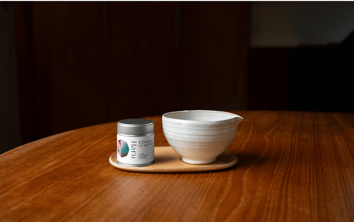 A small white chawan with a pouring spout sits on a round wooden tray next to a silver-lidded tin canister on a wooden table.